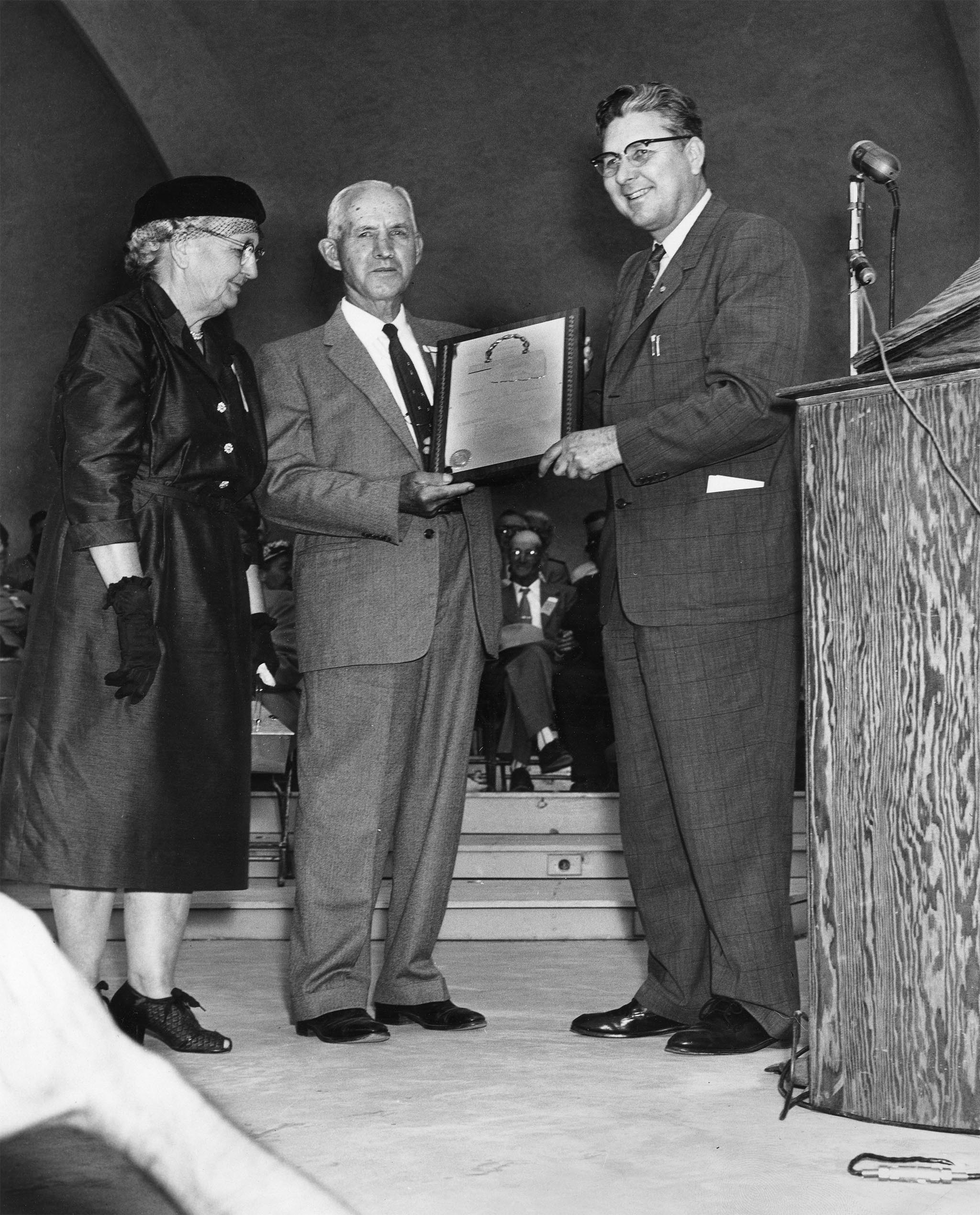 In 1957, the State Fair of Oklahoma asked Farm Bureau to participate in a special Farmers Day at the fair and conduct county contests to select a Golden Anniversary Farm Family. The award was designed to recognize the type of rural people who built Oklahoma in its first 50 years of statehood. The contest was limited to those who had lived on an Oklahoma farm since 1907. A total of 45 county winners were reported by county Farm Bureaus in the contest. Pictured here is Gov. Raymond Gary (right) presenting a gold and silver plaque to the overall winner of the contest, Mr. and Mrs. Fred Graumann, in the bandshell of the State Fair of Oklahoma on September 26, 1957. This was the beginning of a Farm Family award program that continues to today.